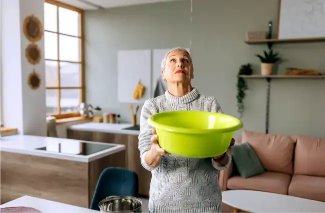 Senior-Woman-Looks-At-The-Ceiling-While-Collecting-Water-Which-Leaks-In-The-Living-Room-At-Home